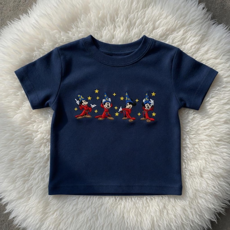 May include: A navy blue short-sleeved t-shirt with an embroidered design. The design features four cartoon characters in red robes and wizard hats, with yellow star accents. The shirt is made of a soft material.