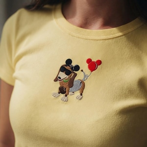 May include: A light yellow t-shirt featuring an embroidered cartoon dachshund wearing sunglasses and a Mickey Mouse-style hat. The dog is holding a red balloon. The design is centered on the chest.