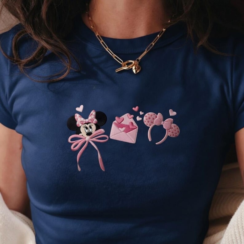 May include: Navy blue t-shirt with embroidered Minnie Mouse and heart designs. The design includes Minnie Mouse, a pink envelope with hearts, and a pink headband with heart-shaped accents. A gold necklace is also visible.