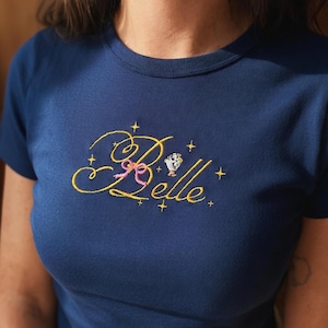 Peut inclure: T-shirt bleu marine avec le nom "Belle" brodé en jaune, orné d'un nœud rose et d'étoiles. Une petite illustration de tasse est également présente. Le t-shirt est de style col rond.