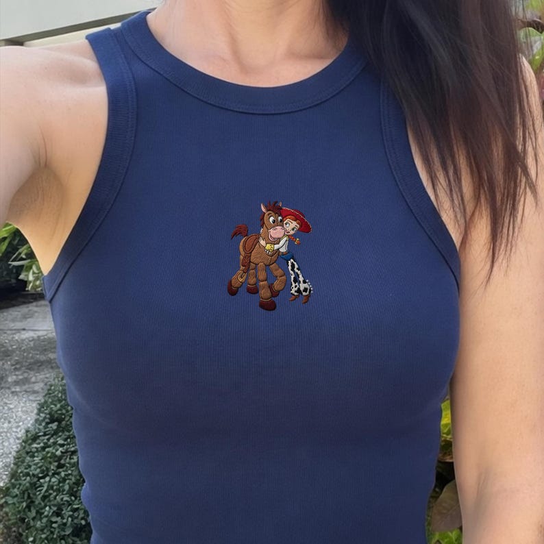 Puede incluir: Camiseta sin mangas de canal&eacute; azul marino con cuello redondo. La camiseta sin mangas presenta un dise&ntilde;o bordado de un caballo de dibujos animados y una vaquera. El dise&ntilde;o es en tonos marrones, rojos, blancos y azules.