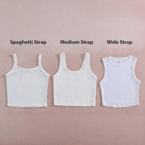 May include: Three white crop tops with different strap styles are displayed against a pink background. The styles are labeled: Spaghetti Strap, Medium Strap, and Wide Strap. The tops are made of a plain, solid white fabric.