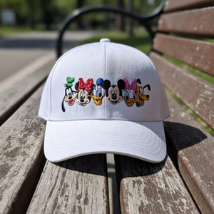 May include: White baseball cap featuring embroidered images of cartoon characters including Goofy, Minnie Mouse, Donald Duck, Mickey Mouse, Daisy Duck, and Pluto. The cap is made of a soft fabric and has a curved brim.