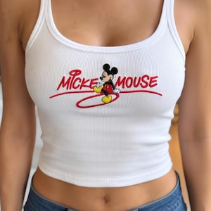 Puede incluir: Camiseta sin mangas blanca acanalada con cuello redondo. La camiseta sin mangas presenta un diseño bordado de Mickey Mouse y las palabras "Mickey Mouse" en escritura roja.