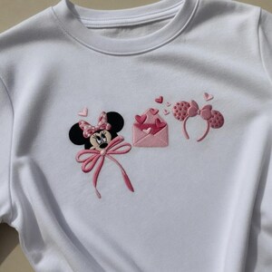May include: White sweatshirt featuring an embroidered design of Minnie Mouse with a pink bow, a pink envelope with hearts, and a pair of pink polka dot ears. The design includes pink hearts and is a cute, whimsical theme.