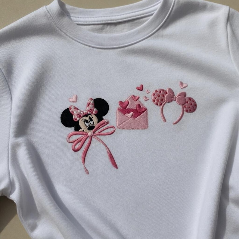 May include: White sweatshirt featuring an embroidered design of Minnie Mouse with a pink bow, a pink envelope with hearts, and a pair of pink polka dot ears. The design includes pink hearts and is a cute, whimsical theme.