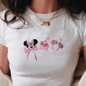 May include: White t-shirt featuring a Minnie Mouse design with a pink bow, hearts, and an envelope. The design includes a Minnie Mouse head, an envelope with hearts, and a headband with Minnie Mouse ears.