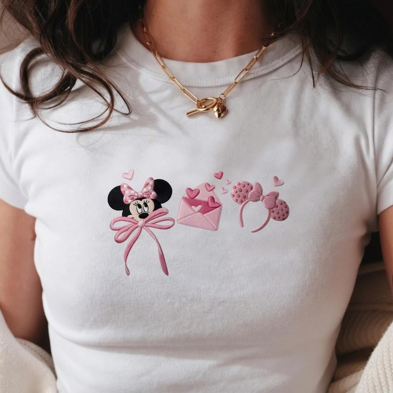 May include: White t-shirt featuring a Minnie Mouse design with a pink bow, hearts, and an envelope. The design includes a Minnie Mouse head, an envelope with hearts, and a headband with Minnie Mouse ears.