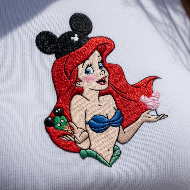 May include: Embroidered patch featuring a cartoon mermaid with red hair, blue seashell bra, and a black Mickey Mouse ear hat. The mermaid is holding a pink flower and a green ice cream cone. The patch is on a white fabric.