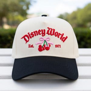 May include: A cream-colored baseball cap with a black brim. The cap features the embroidered text "Disney World" in red, along with a cherry graphic and the year "1971". The cap is a stylish accessory.