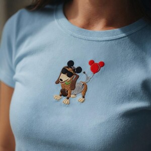 May include: A light blue t-shirt featuring an embroidered cartoon dog wearing sunglasses and a green collar, holding a red balloon. The dog has a skeletal design on its body and mouse ears.