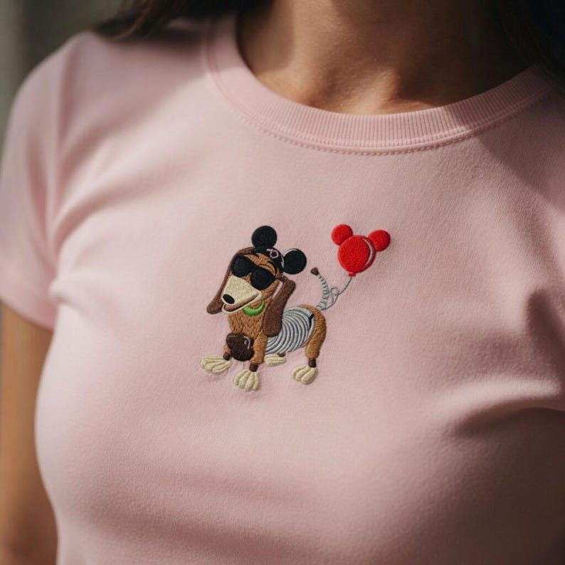 May include: A light pink t-shirt featuring an embroidered cartoon dachshund wearing sunglasses and a Mickey Mouse-style hat. The dog is holding a red balloon. The dog has a brown body, tan paws, and a green collar.