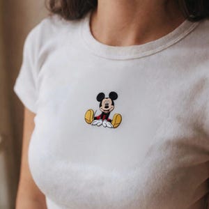 K&ouml;nnte beinhalten: Wei&szlig;es T-Shirt mit einem gestickten Mickey Mouse-Motiv. Die Zeichentrickfigur ist sitzend dargestellt und tr&auml;gt rote Shorts, gelbe Schuhe und wei&szlig;e Handschuhe. Das Shirt hat einen Rundhalsausschnitt und kurze &Auml;rmel.