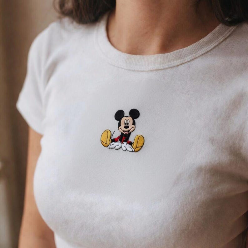K&ouml;nnte beinhalten: Wei&szlig;es T-Shirt mit einem gestickten Mickey Mouse-Motiv. Die Zeichentrickfigur ist sitzend dargestellt und tr&auml;gt rote Shorts, gelbe Schuhe und wei&szlig;e Handschuhe. Das Shirt hat einen Rundhalsausschnitt und kurze &Auml;rmel.