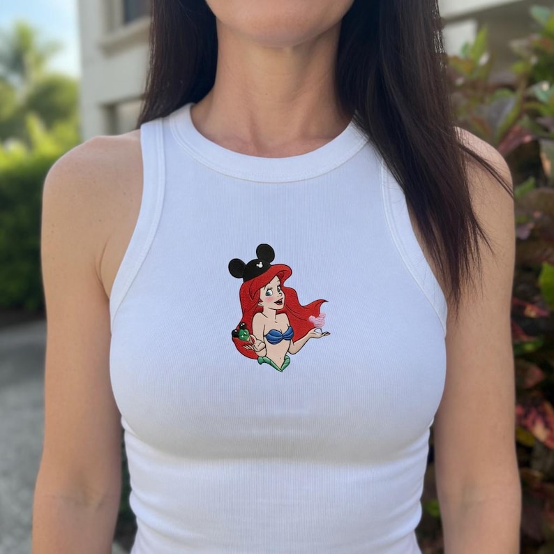 May include: White ribbed tank top featuring a colorful cartoon graphic of Ariel from The Little Mermaid wearing a black Mickey Mouse ear hat. The graphic includes Ariel's red hair, blue seashell bra, and green tail.