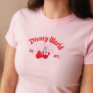 May include: Light pink t-shirt with a crew neck. The shirt features an embroidered design of cherries and the words "Disney World" in red script, with "Est. 1971" below. The cherries have green stems and a pink bow.