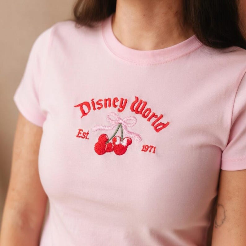 May include: Light pink t-shirt with a crew neck. The shirt features an embroidered design of cherries and the words "Disney World" in red script, with "Est. 1971" below. The cherries have green stems and a pink bow.