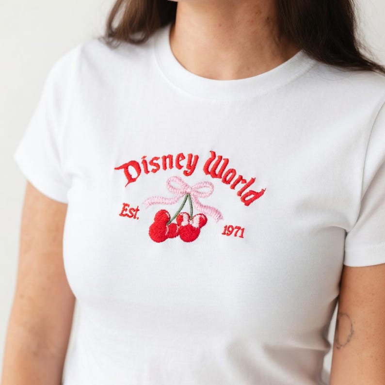 May include: White t-shirt with a red embroidered "Disney World" logo, featuring cherries and a pink bow. The text "Est. 1971" is also embroidered on the shirt. The shirt is a crew neck style.