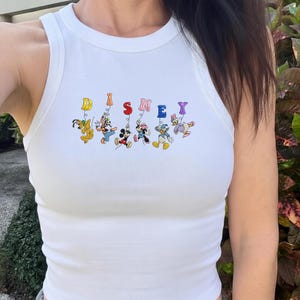 May include: White tank top with the word "DISNEY" in colorful letters. The letters are decorated with cartoon characters including Pluto, Mickey Mouse, Minnie Mouse, Donald Duck, and Daisy Duck.