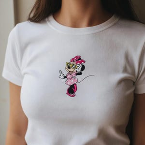May include: White t-shirt featuring a cartoon Minnie Mouse design. Minnie is depicted in a pink dress with white polka dots, a matching bow, and yellow sunglasses. The design is centered on the chest.