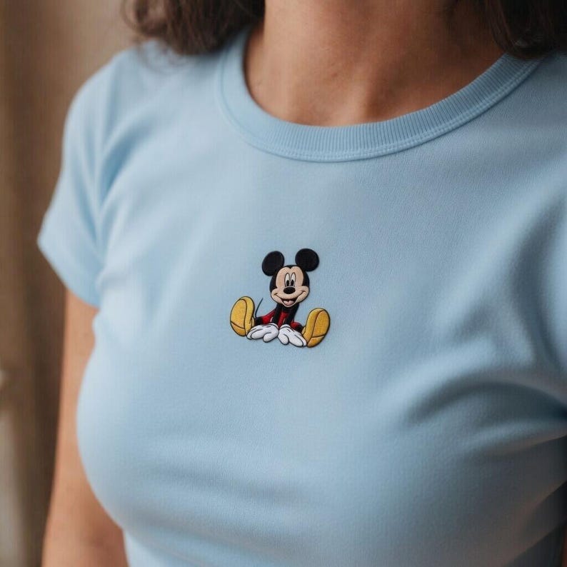 K&ouml;nnte beinhalten: Hellblaues T-Shirt mit einer kleinen Mickey Mouse-Stickerei. Die Cartoonfigur ist sitzend dargestellt und tr&auml;gt rote Shorts, gelbe Schuhe und wei&szlig;e Handschuhe. Das Shirt hat einen Rundhalsausschnitt und kurze &Auml;rmel.