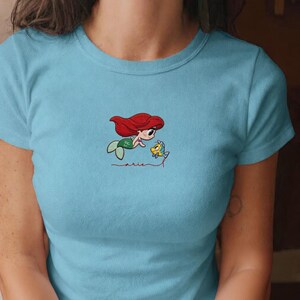 May include: Pale blue t-shirt with an embroidered design of a cartoon mermaid with red hair and a green tail, alongside a yellow fish. The word "arie" is embroidered in a cursive font below the characters.