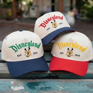 Puede incluir: Tres gorras de béisbol con la palabra "Disneyland" bordada en verde, rojo y amarillo. Cada gorra tiene una corona blanca, una visera de color y un gráfico de Pluto. Las gorras también presentan el texto "EST 1955".