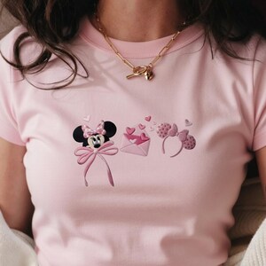 May include: A light pink t-shirt featuring embroidered designs of Minnie Mouse, an envelope with hearts, and a pair of Minnie Mouse ears. The shirt has a round neckline and short sleeves. A gold necklace is worn around the neck.