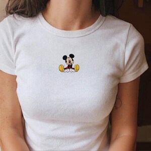 K&ouml;nnte beinhalten: Wei&szlig;es T-Shirt mit einem kleinen, gestickten Mickey Mouse-Motiv. Die Zeichentrickfigur ist sitzend dargestellt und tr&auml;gt rote Shorts, gelbe Schuhe sowie einen schwarzen K&ouml;rper und Kopf. Das Shirt hat einen Rundhalsausschnitt und kurze &Auml;rmel.