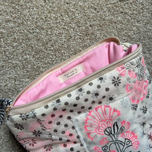 May include: Open beige zippered pouch with a pink interior. The exterior features a floral and polka dot pattern in black and pink. The zipper is beige, and the pull is black and white. The label inside says "hand made by..."