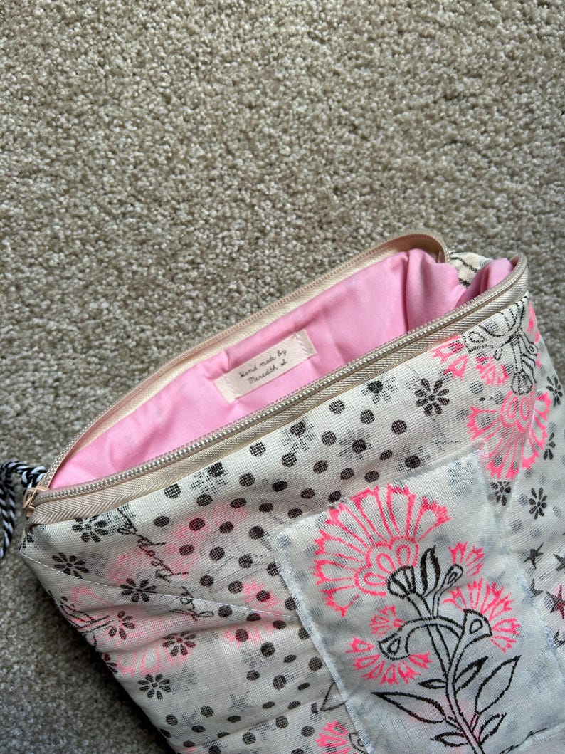 May include: Open beige zippered pouch with a pink interior. The exterior features a floral and polka dot pattern in black and pink. The zipper is beige, and the pull is black and white. The label inside says "hand made by..."
