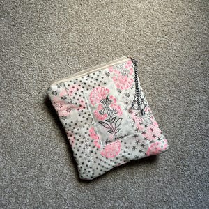 May include: A quilted clutch with a floral pattern in pink and white, accented with black polka dots and stars. The text "free people" is visible. The clutch has a zipper closure and a black and white rope handle.
