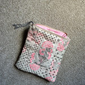May include: A quilted pouch with a floral design in pink and black on a white background. The pouch has a zipper closure and a black and white cord loop. The interior is pink.