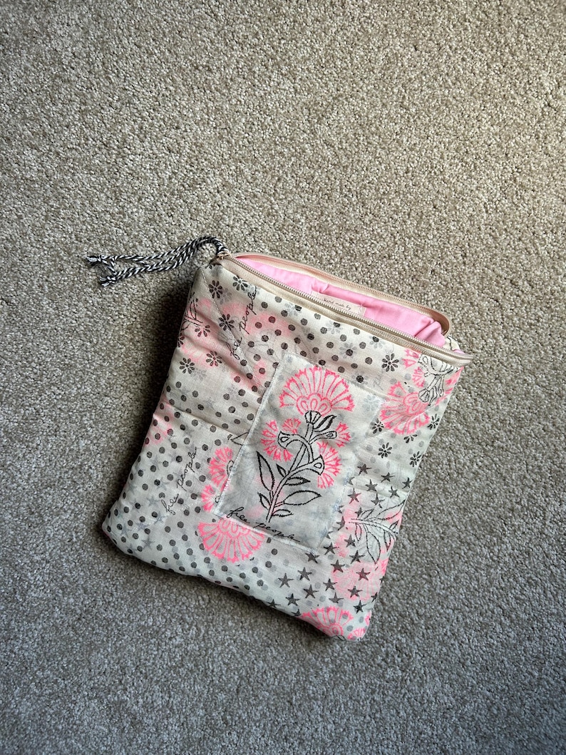 May include: A quilted pouch with a floral design in pink and black on a white background. The pouch has a zipper closure and a black and white cord loop. The interior is pink.