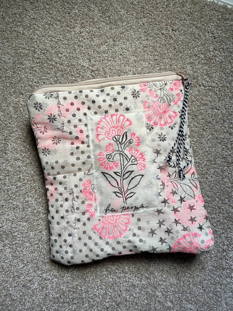 May include: A beige and pink floral patterned pouch with a zipper closure. The pouch features a floral design with the text "free people". The pouch has a black and white tassel attached to the zipper.