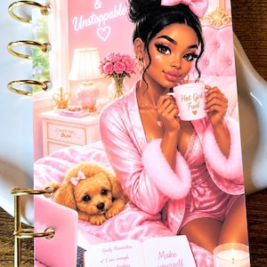 May include: A spiral-bound notebook featuring an illustration of a person in a pink robe holding a mug labeled Hot Girl Fuel, with a puppy, a laptop, and an open book with the text Make yourself proud and Daily Reminders written inside.