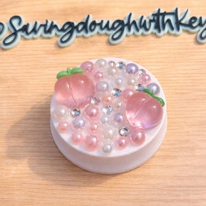 May include: A white, round decorative object adorned with pink and white pearls, clear rhinestones, and two translucent pink peach-shaped embellishments with green leaves. The text "@swingdoughwithkey" is above the object.