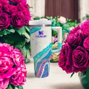 May include: A white Stanley tumbler featuring a glittery, multi-colored geode-inspired design with swirling layers of teal, blue, and purple. The cup includes a sturdy handle, a white lid, and the Stanley logo printed in dark blue on the front.