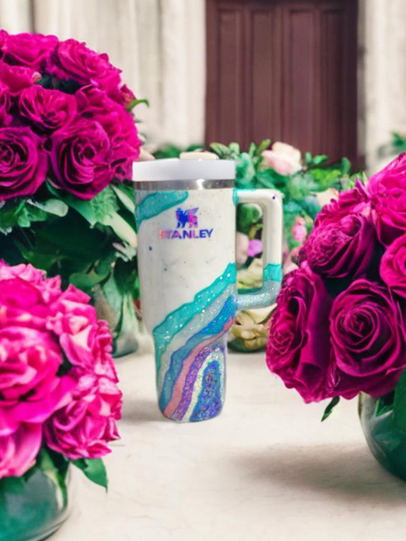 May include: A white Stanley tumbler featuring a glittery, multi-colored geode-inspired design with swirling layers of teal, blue, and purple. The cup includes a sturdy handle, a white lid, and the Stanley logo printed in dark blue on the front.