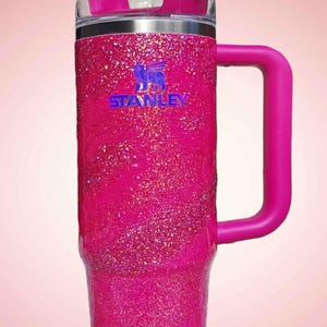 Pode incluir: Um copo Stanley rosa brilhante com uma al&ccedil;a e um canudo. O copo tem o logotipo Stanley em azul. A tampa e a al&ccedil;a s&atilde;o de cor rosa s&oacute;lida. O copo foi projetado para manter as bebidas frias ou quentes.