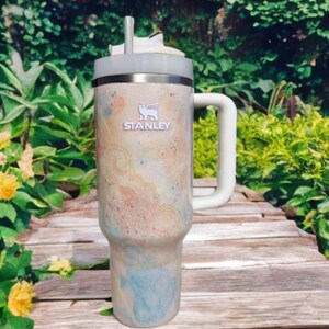 Puede incluir: Un vaso t&eacute;rmico de la marca Stanley con asa. El vaso presenta un dise&ntilde;o en crema, azul y melocot&oacute;n. Tiene un asa blanca y una tapa gris con pajita. El logotipo de Stanley es visible en la parte delantera.