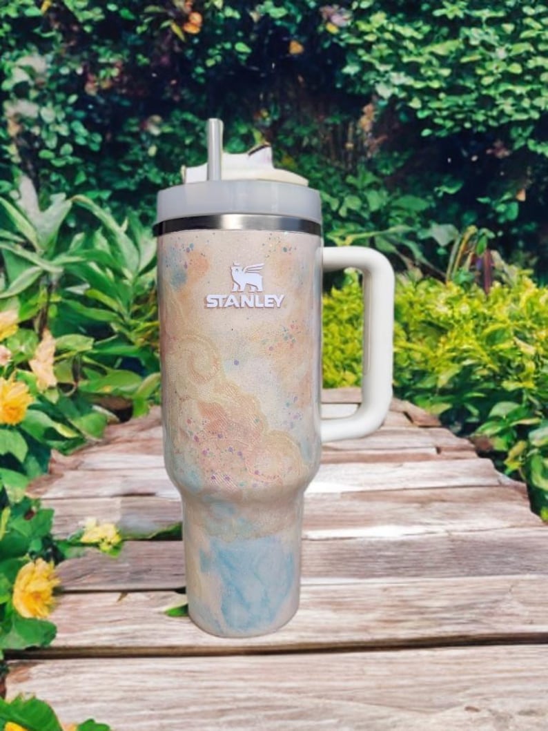 Puede incluir: Un vaso t&eacute;rmico de la marca Stanley con asa. El vaso presenta un dise&ntilde;o en crema, azul y melocot&oacute;n. Tiene un asa blanca y una tapa gris con pajita. El logotipo de Stanley es visible en la parte delantera.