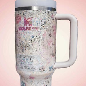 May include: A Stanley tumbler with a white handle and lid, featuring a floral and glitter design. The tumbler has the Stanley logo and the text "FOCUS on the good". The tumbler is designed for cold beverages.