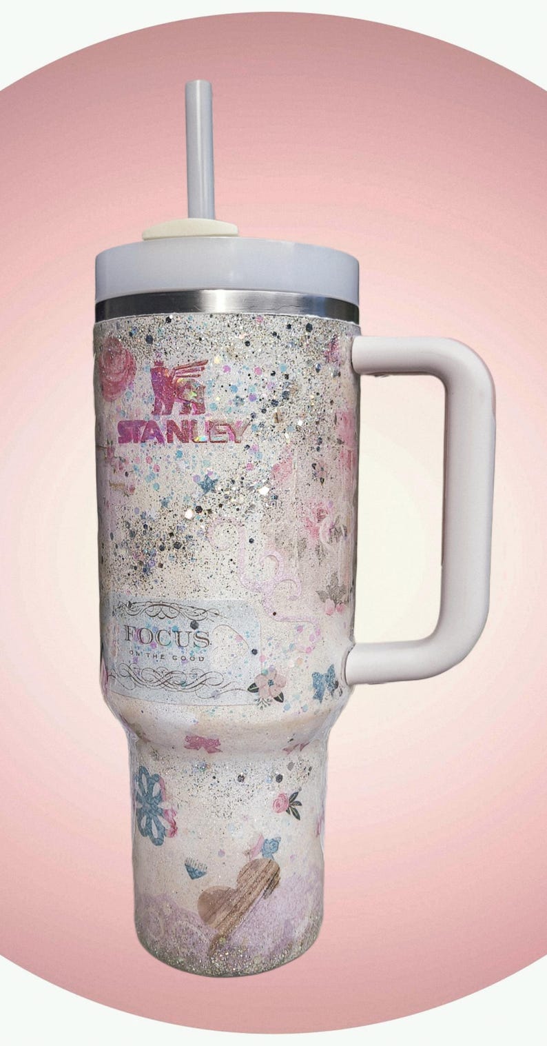 May include: A Stanley tumbler with a white handle and lid, featuring a floral and glitter design. The tumbler has the Stanley logo and the text "FOCUS on the good". The tumbler is designed for cold beverages.