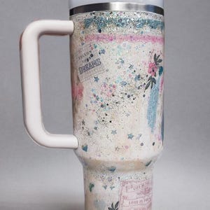May include: A white and silver insulated tumbler with a handle, lid, and straw. The tumbler is decorated with glitter, floral accents, and the words "Follow Your Dreams" and "Love is for".