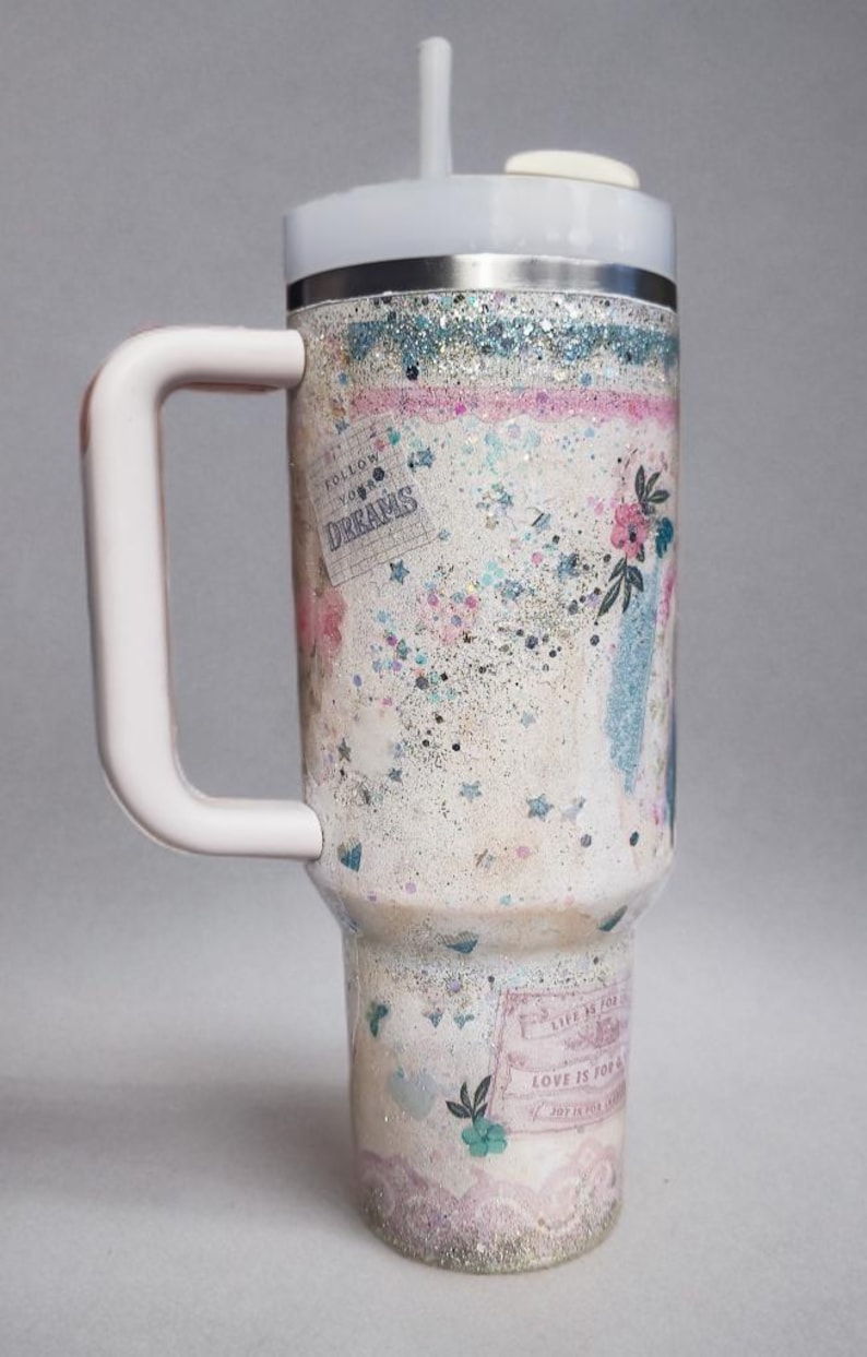 May include: A white and silver insulated tumbler with a handle, lid, and straw. The tumbler is decorated with glitter, floral accents, and the words "Follow Your Dreams" and "Love is for".