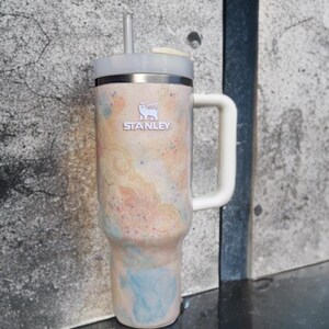 Puede incluir: Un vaso t&eacute;rmico Stanley con asa y pajita. El vaso presenta un dise&ntilde;o abstracto colorido en tonos melocot&oacute;n, azul y rosa. El logotipo de Stanley es visible en la parte delantera.