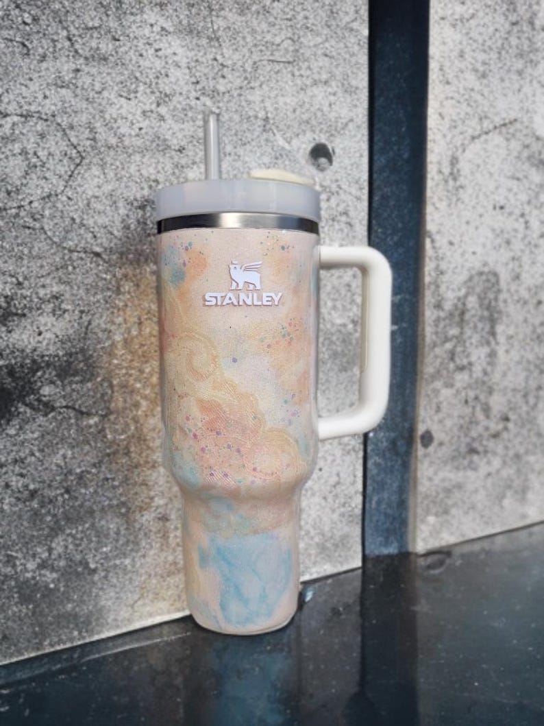 Puede incluir: Un vaso t&eacute;rmico Stanley con asa y pajita. El vaso presenta un dise&ntilde;o abstracto colorido en tonos melocot&oacute;n, azul y rosa. El logotipo de Stanley es visible en la parte delantera.
