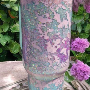 Puede incluir: Un vaso cil&iacute;ndrico con un acabado de m&uacute;ltiples capas con parches de tonos rosa pastel, azul claro y morado. La superficie est&aacute; cubierta con una textura de purpurina fina y brillante, y el vaso est&aacute; rematado con una tapa de color azul claro.
