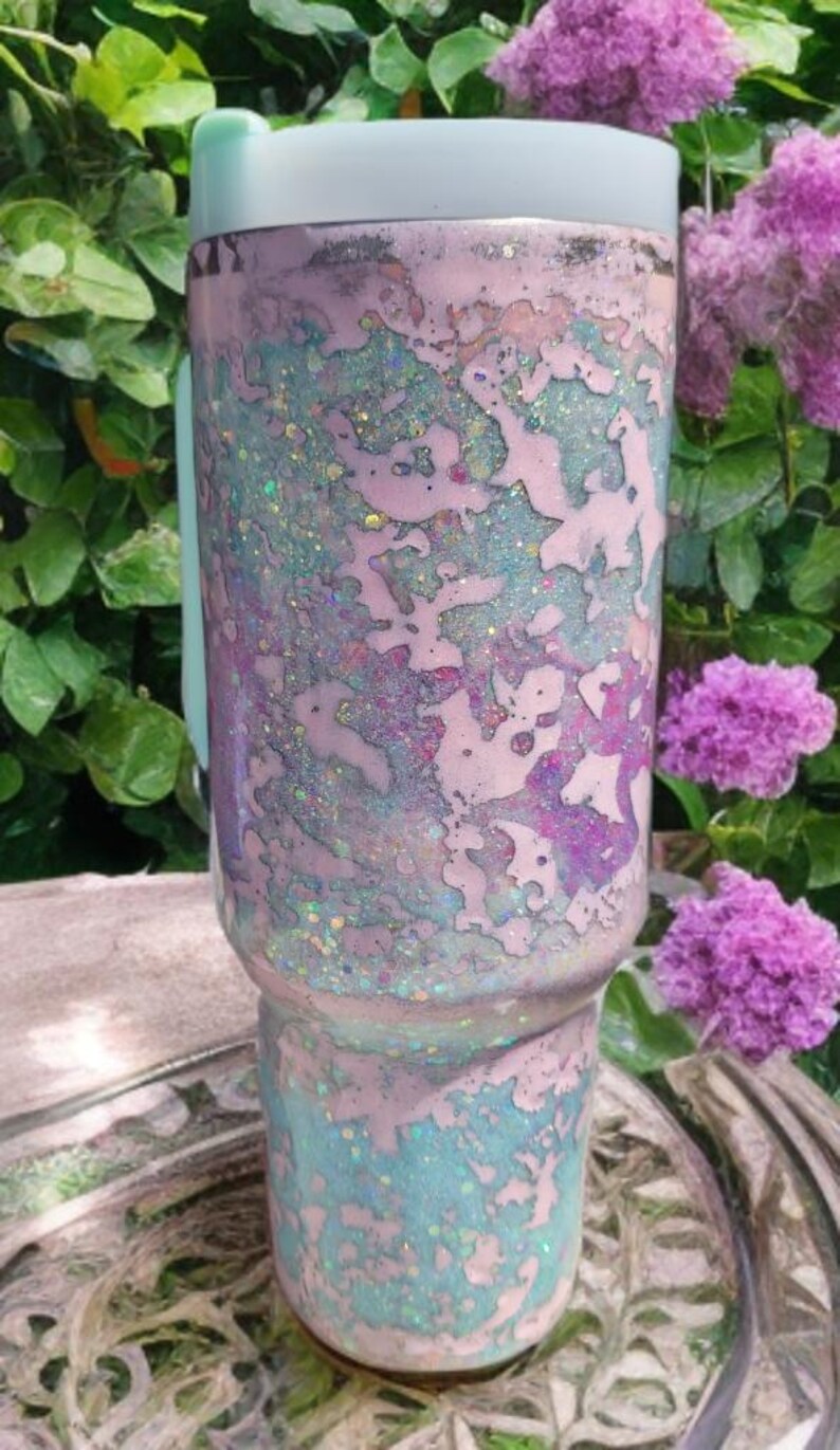 Puede incluir: Un vaso cil&iacute;ndrico con un acabado de m&uacute;ltiples capas con parches de tonos rosa pastel, azul claro y morado. La superficie est&aacute; cubierta con una textura de purpurina fina y brillante, y el vaso est&aacute; rematado con una tapa de color azul claro.
